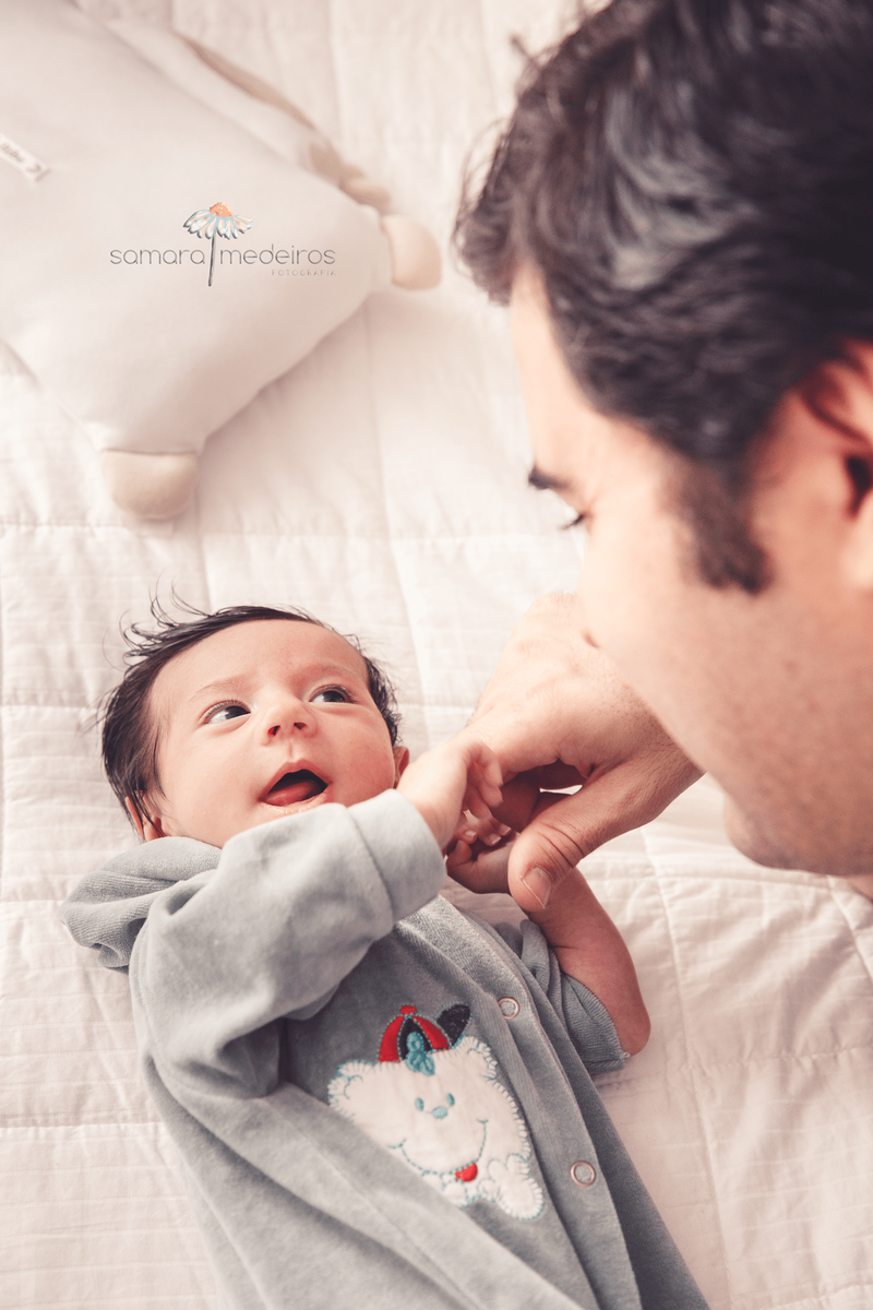 Bebê recém nascido deitado na cama dos pais, segurando o dedo do seu pai e sorrindo.