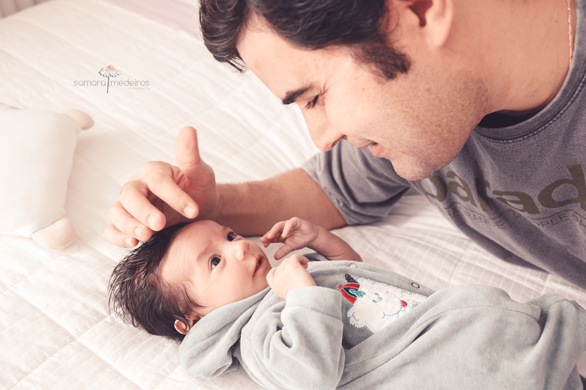 Pai e seu filho recém-nascido deitados na cama e se olhando, durante ensaio newborn lifestyle.