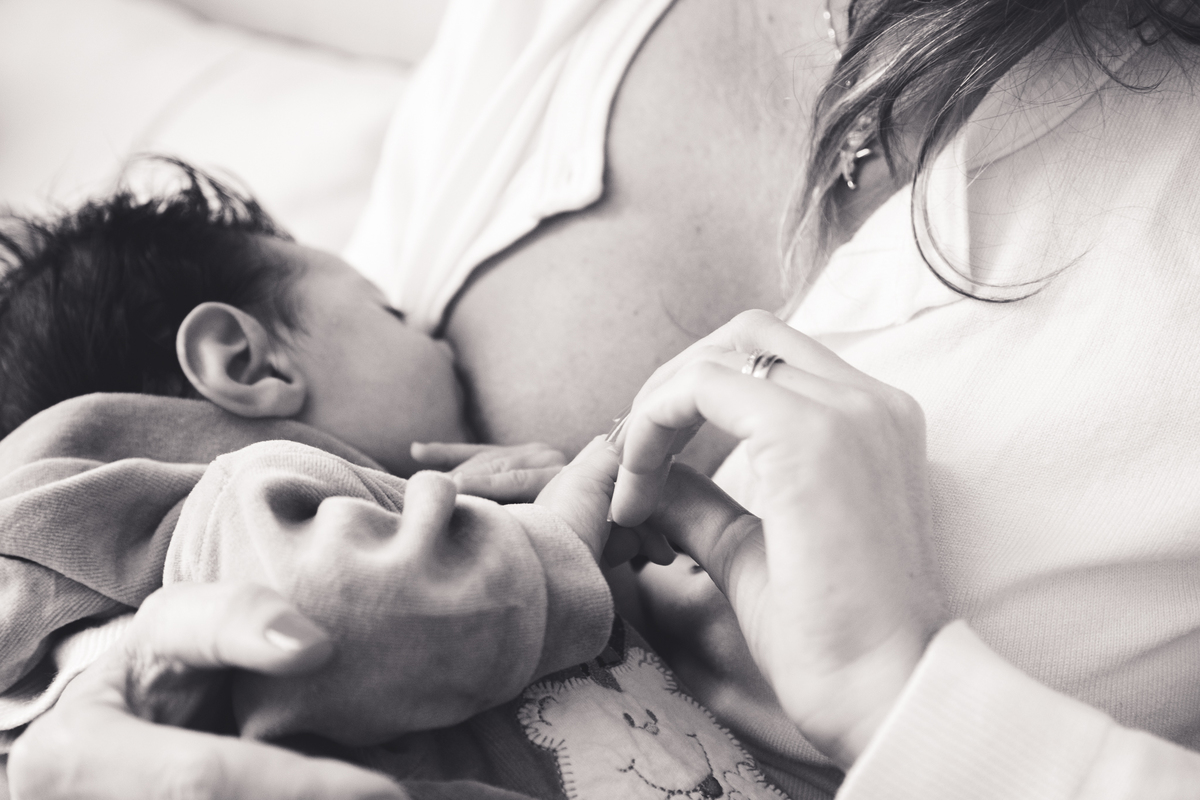 Foto em preto e branco do bebê recém nascido mamando, com sua mãe segurando sua mãozinha, fazendo carinho.