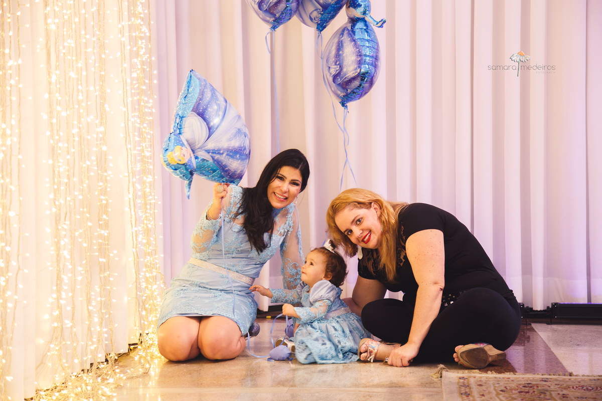 Duas adultas sentadas no chão ao lado de uma criança de um ano, brincando com um balão, durante sua festa de aniversário.