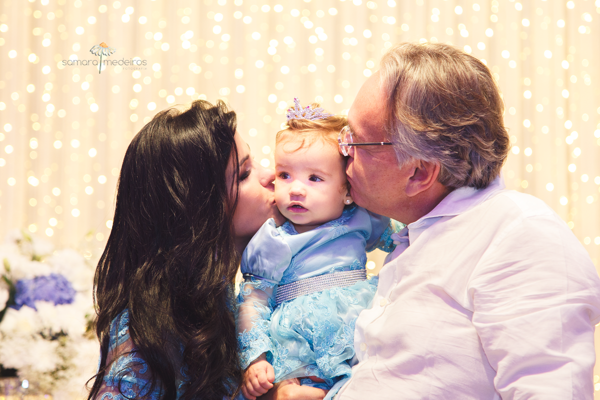 Bebê de 1 ano ganhando beijo dos seus pais, um em cada bochecha, durante sua festa de aniversário.