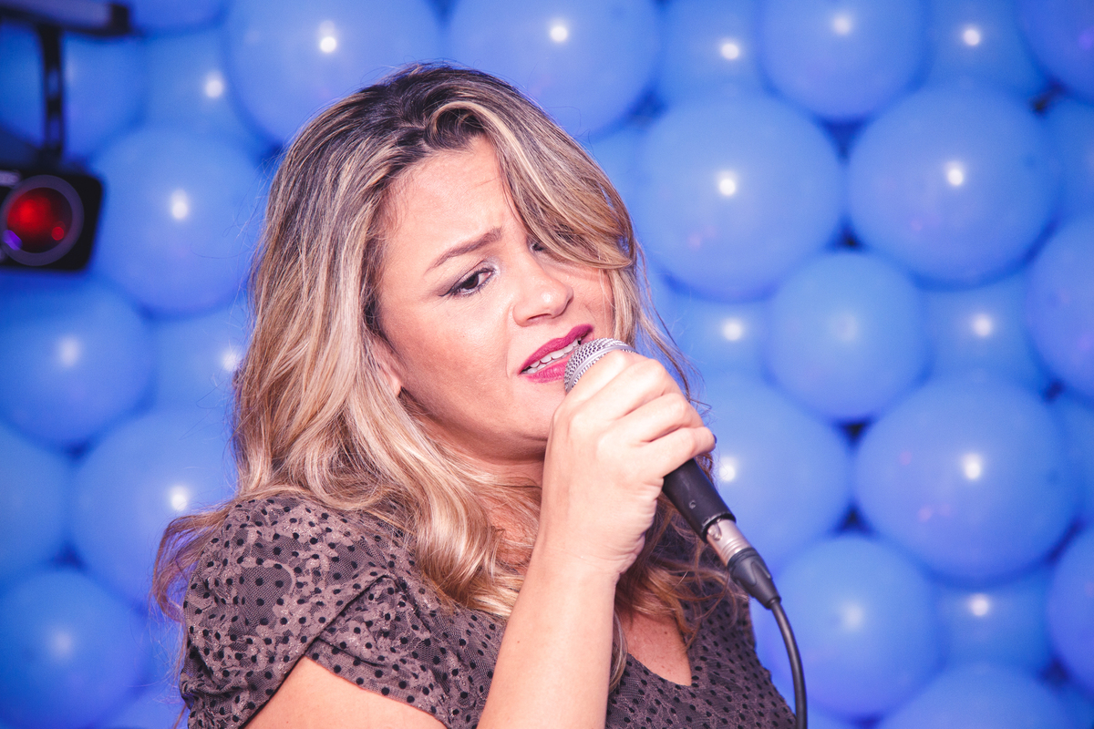 Mulher cantando com um microfone à boca e um fundo de balões azuis.