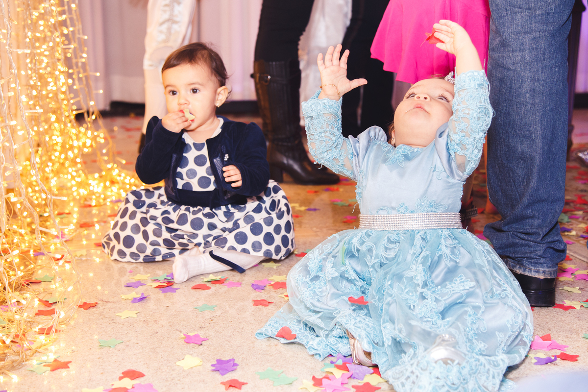 Duas crianças sentadas no chão em festa de aniversário, após o parabéns, brincando com os papéis picados que caíram após o parabéns.