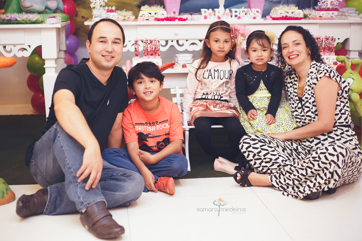 Família junta em frente à mesa do bolo, pai, mãe e três crianças, todos sentados no chão ou no banquinho em frente à mesa.