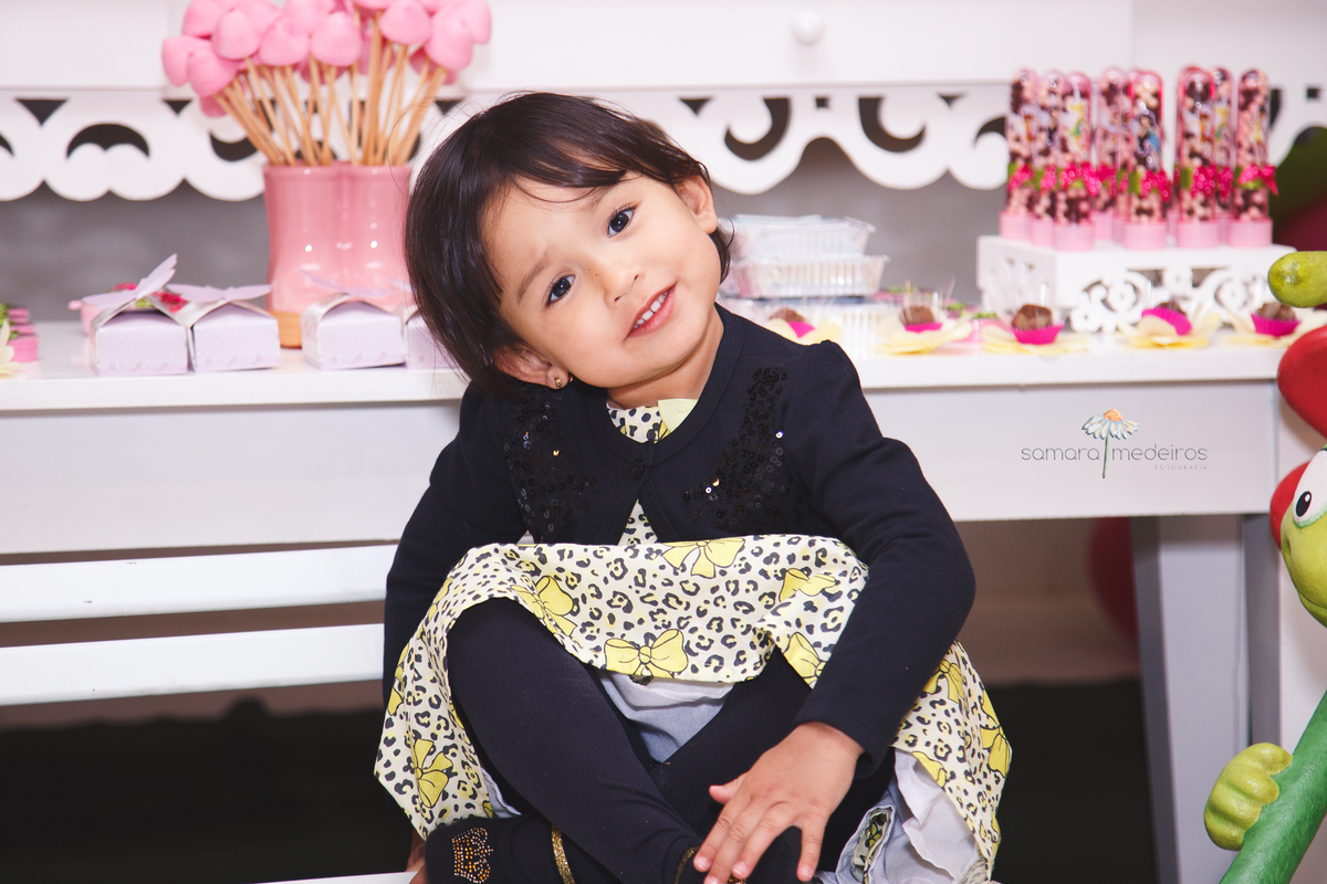 Menina de 3 anos posando para foto com uma expressão singela, sentada em um banquinho de praça infantil, posicionado em frente à mesa do bolo do seu aniversário.