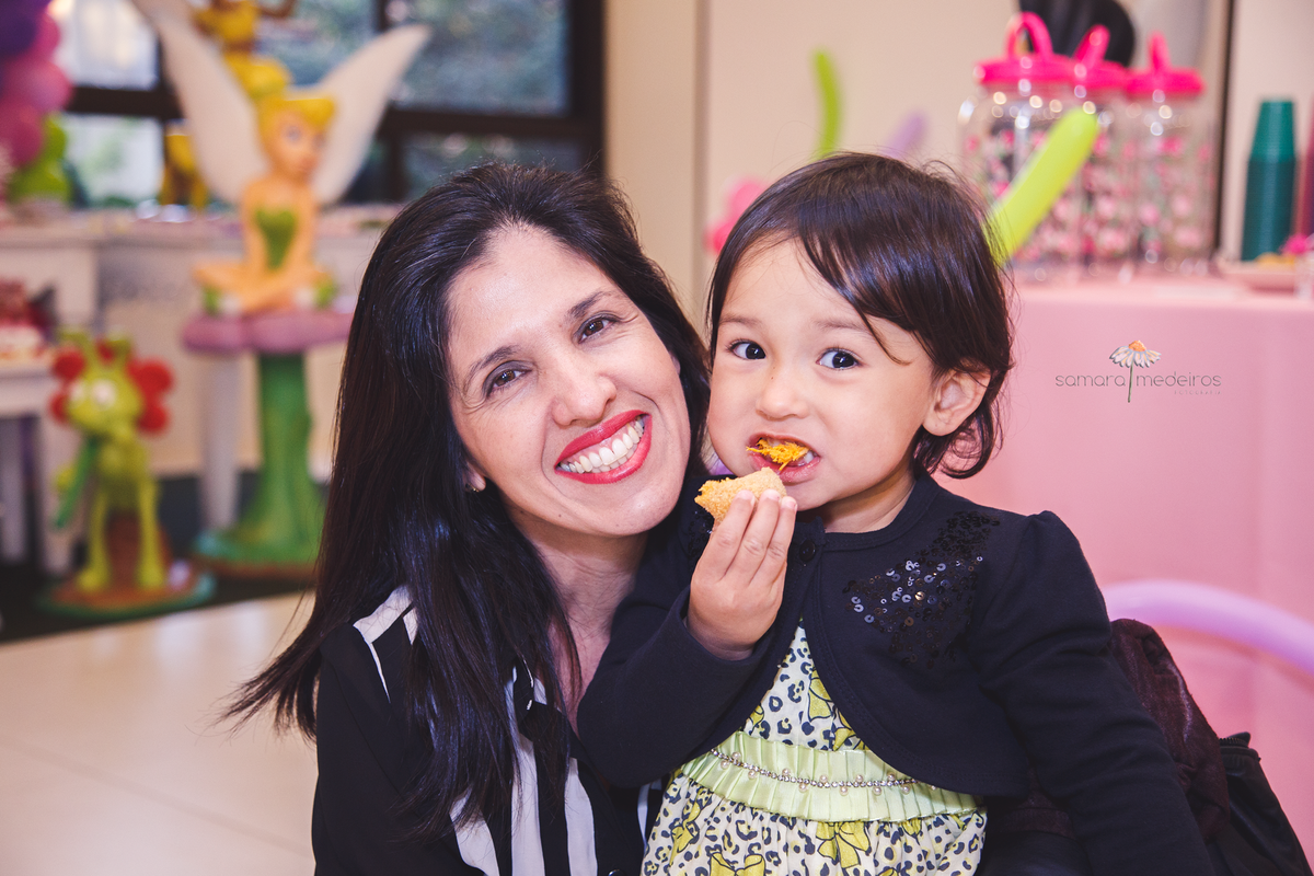 Criança de três anos posando para foto com sua tia, enquanto come uma coxinha e sorri ao mesmo tempo.