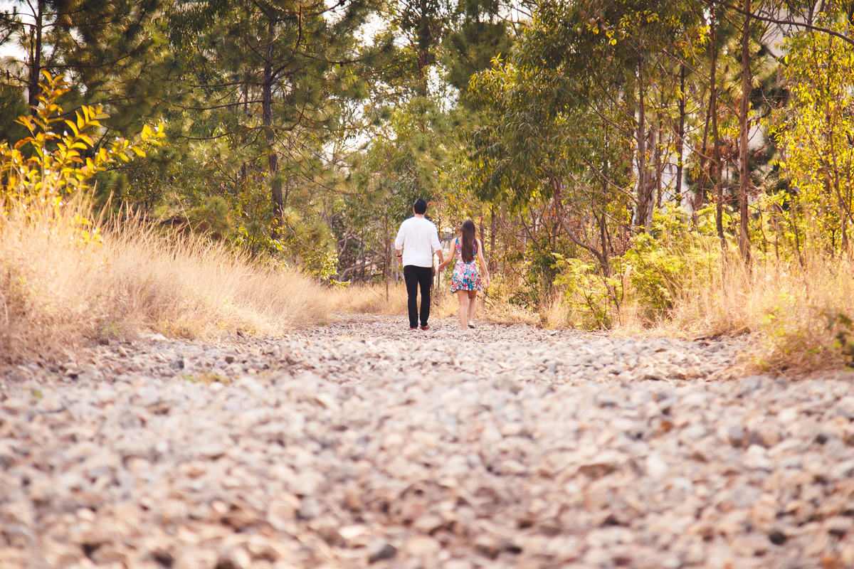 Casal andando de mãos dadas, ao longe, em uma estrada ladeada por árvores e coberta de pedrinhas.