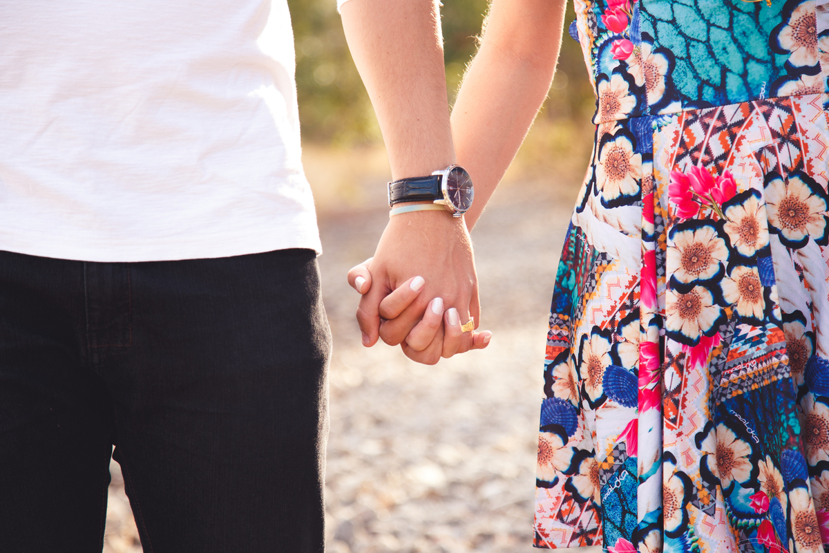 Detalhe do casal de mãos dadas, foco na mão deles, com dedos entrelaçados e parte do corpo de cada um.