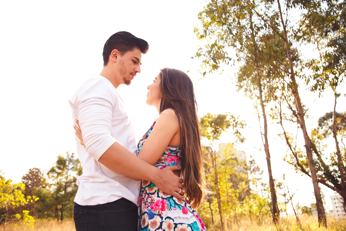 Casal abraçado, um de frente para o outro, se olhando nos olhos, com o sol e árvores ao fundo.