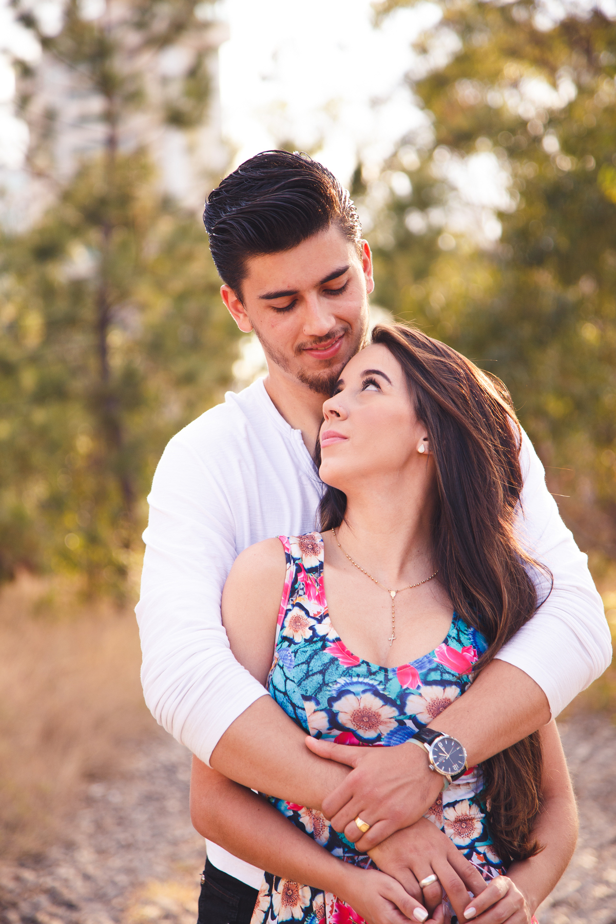 Casal abraçado, a moça de costas para o rapaz, apoiada no peito dele e se olhando. árvores ao fundo.