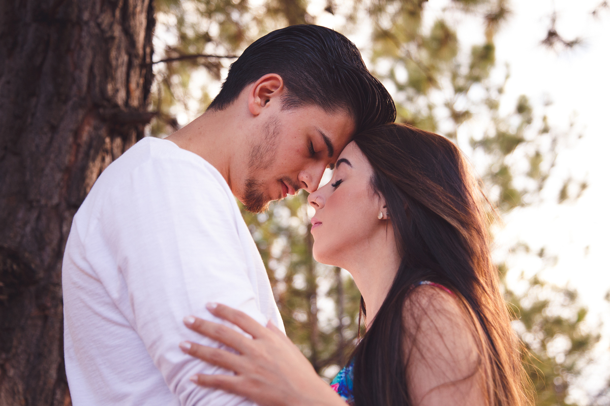 Casal ao lado de uma árvore, se abraçando, com as testas encostadas, de olhos fechados, durante fotos de uma sessão casal.