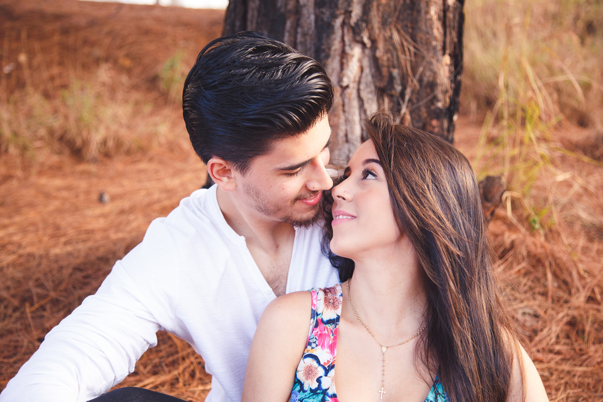Casal sentado, o rapaz apoiado em uma árvore e a moça apoiada no peito dele, se olhando nos olhos e sorrindo.