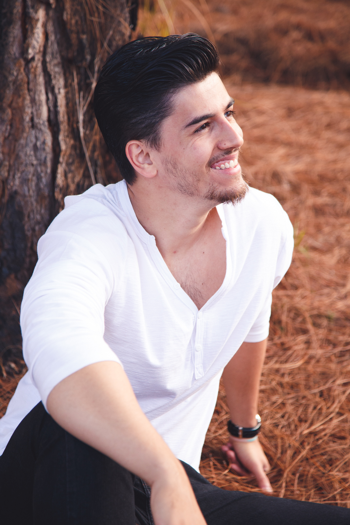 Rapaz sentado apoiado em uma árvore olhando para a namorada, que não aparece na foto, com uma expressão sorridente.