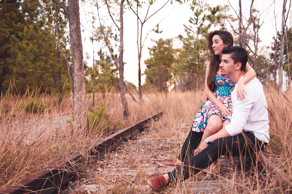 Casal sentado em um trilho de trem desativado, a moça no colo do rapaz, ambos com expressão pensativa observando à frente.