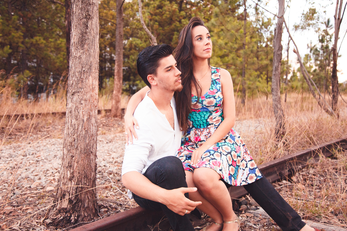 Casal sentado em um trilho de trem desativado, a moça no colo do rapaz, olhando distraidamente à frente.