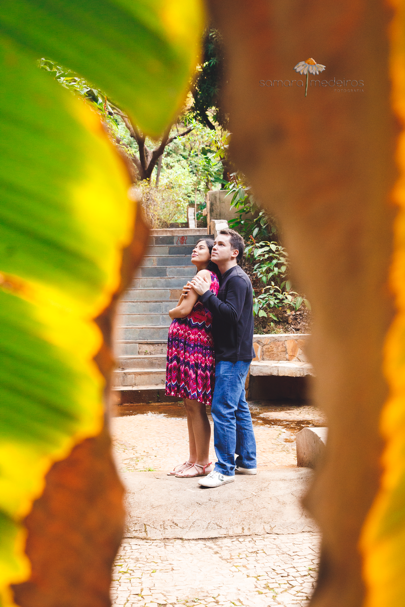 Mãe gestante e pai de pé abraçados, com expressão pensativa, sendo vistos entre as folhas de uma bananeira.