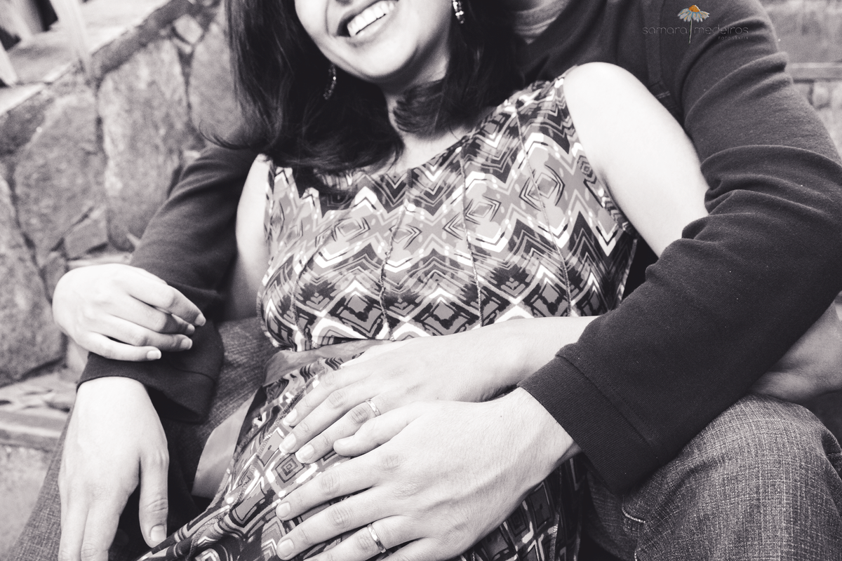 Foto em preto e branco de um casal, sendo que a mulher está gestante, sorrindo, ambos com a mão sobre a barriga.