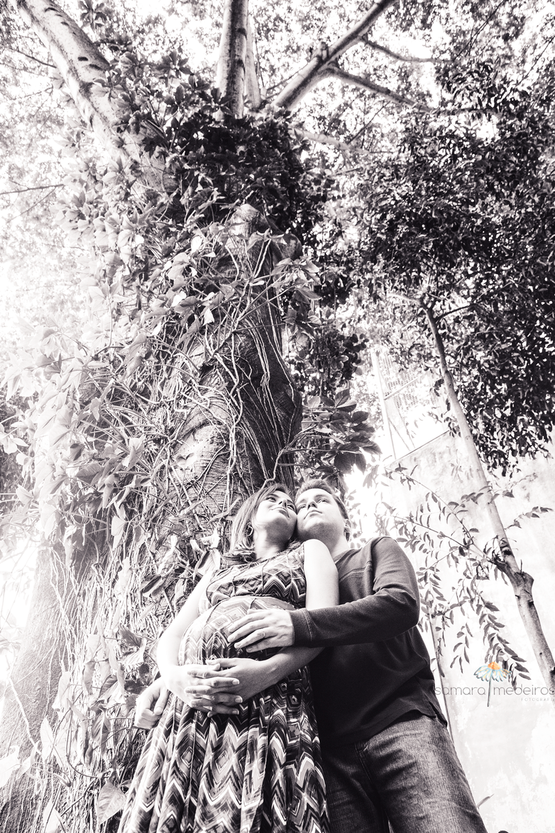 Foto em preto e branco de futuros pais de pé em frente a uma árvore enorme, olhando distraídos para frente, mãos apoiadas na barriga da mãe.