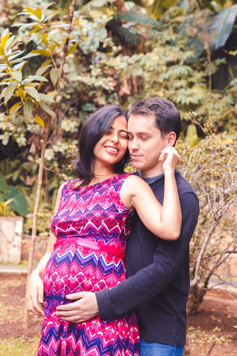 Marido e esposa abraçados, com um leve sorriso, olhos fechados e expressão serena, enquanto apoiam as mãos na barriga da gestante.