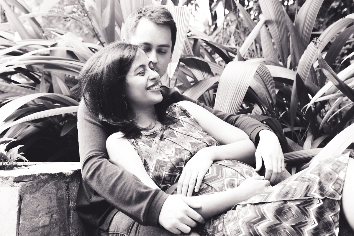 Foto em preto e branco de uma mãe gestante recostada no marido, com as mãos apoiadas na barriga, sorrindo.