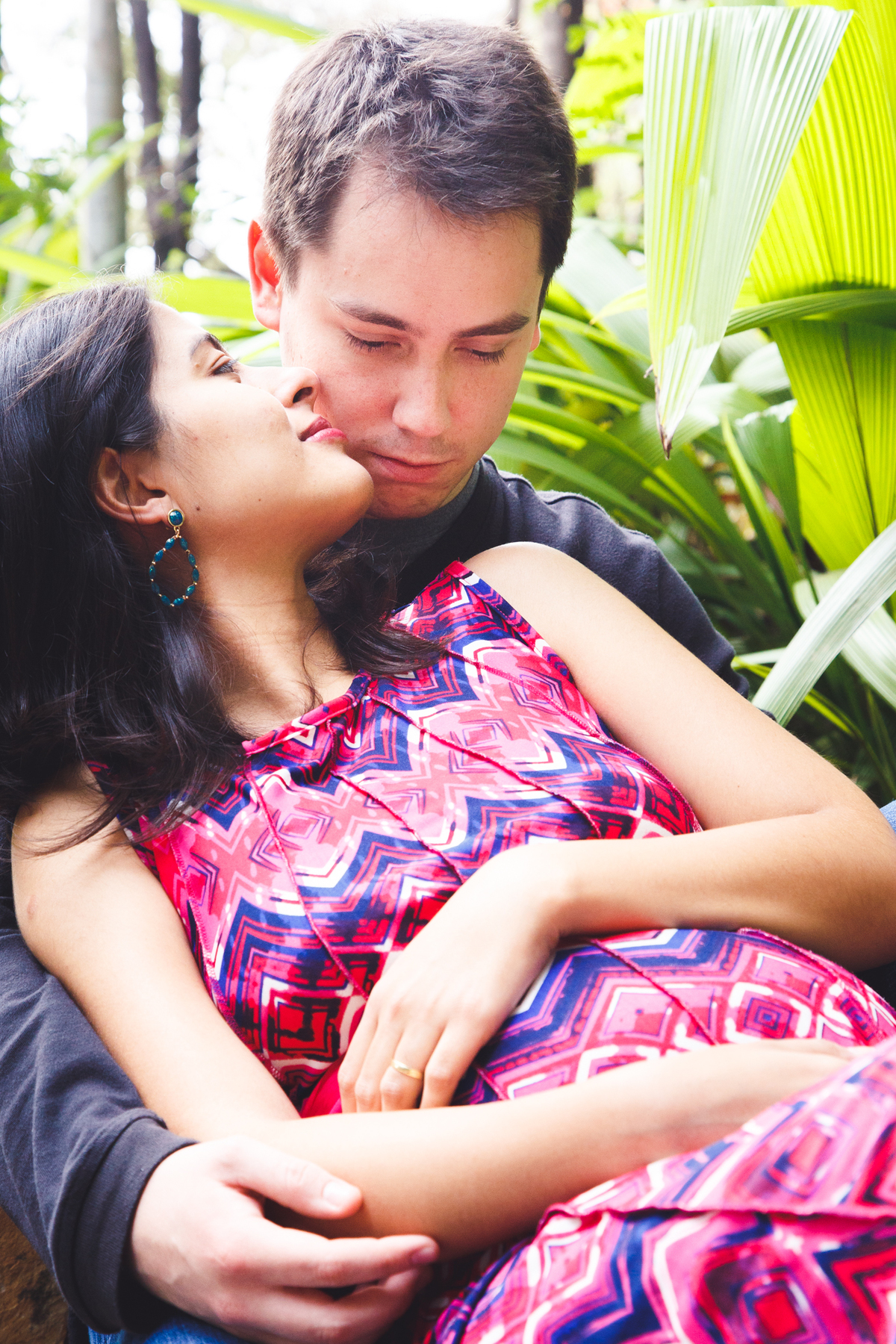 Mãe grávida sentada em um banco apoiada no marido atrás dela. Ela apoia a mão na barriga e olha para e ele.