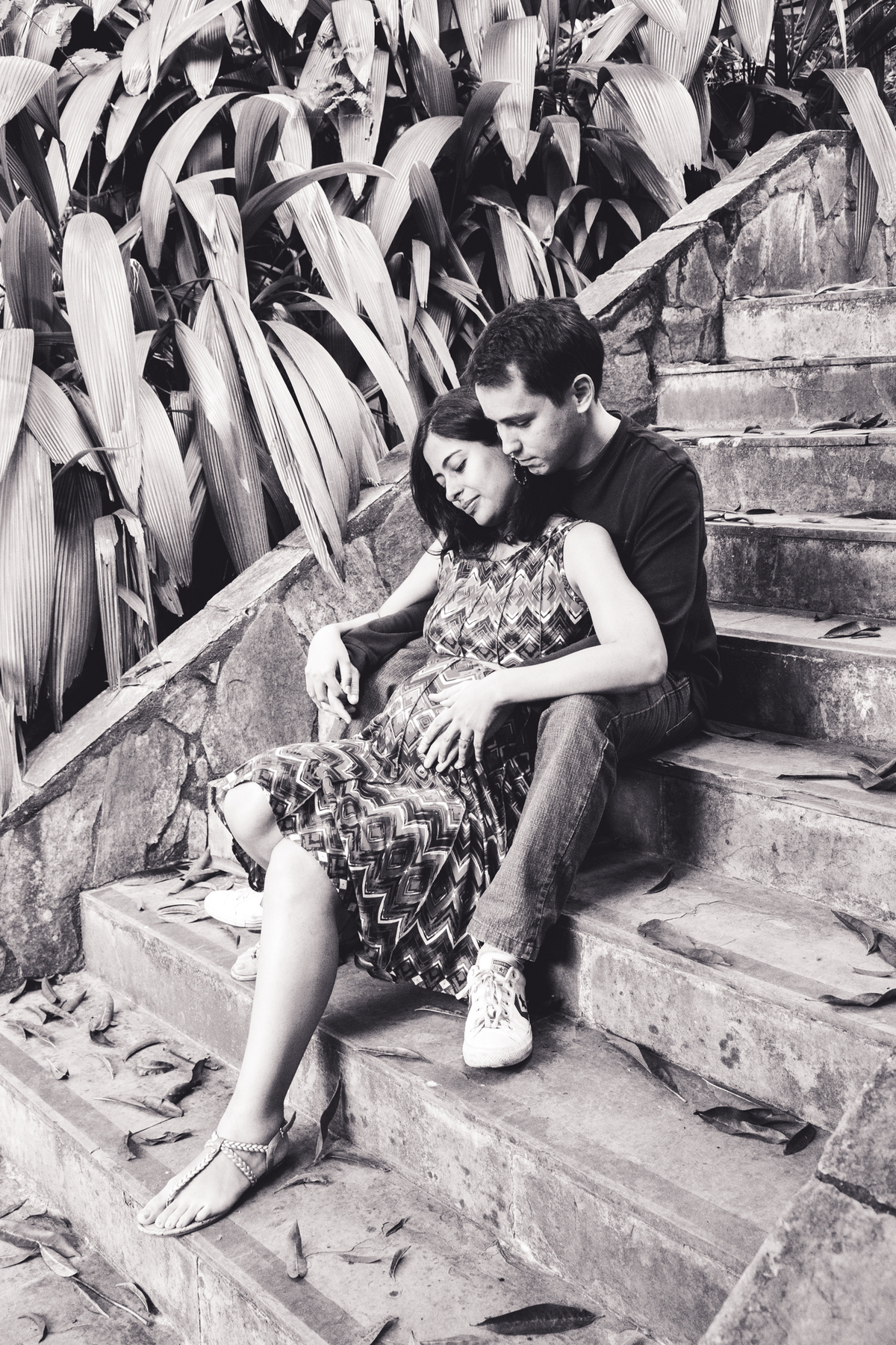 Foto em preto e branco de uma mulher grávida sentada em uma escada apoiada no marido, ambos sérios, com expressão pensativa, observando a barriga.