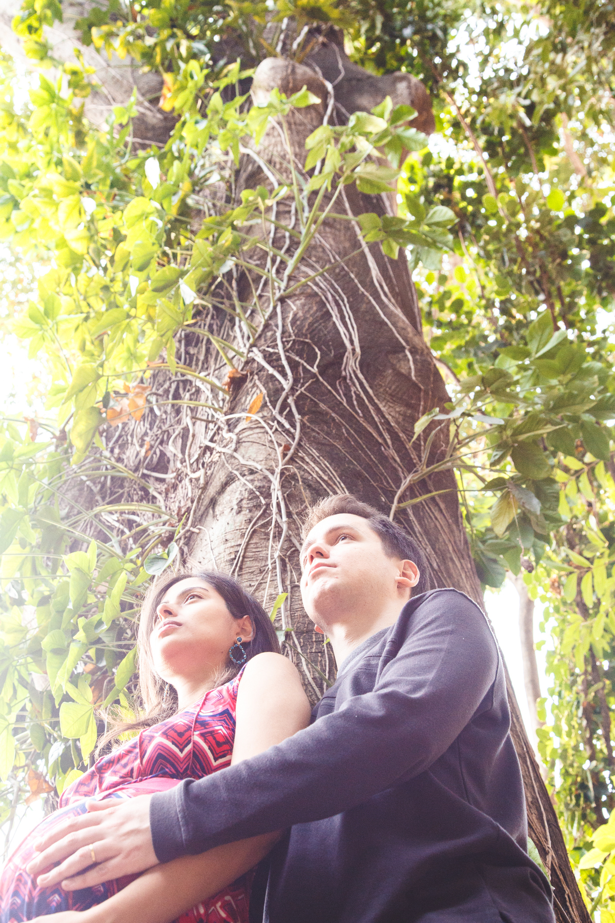 Futuros pais em cima de uma plataforma, de costas para uma árvore grande enquanto olham distraídos para frente.