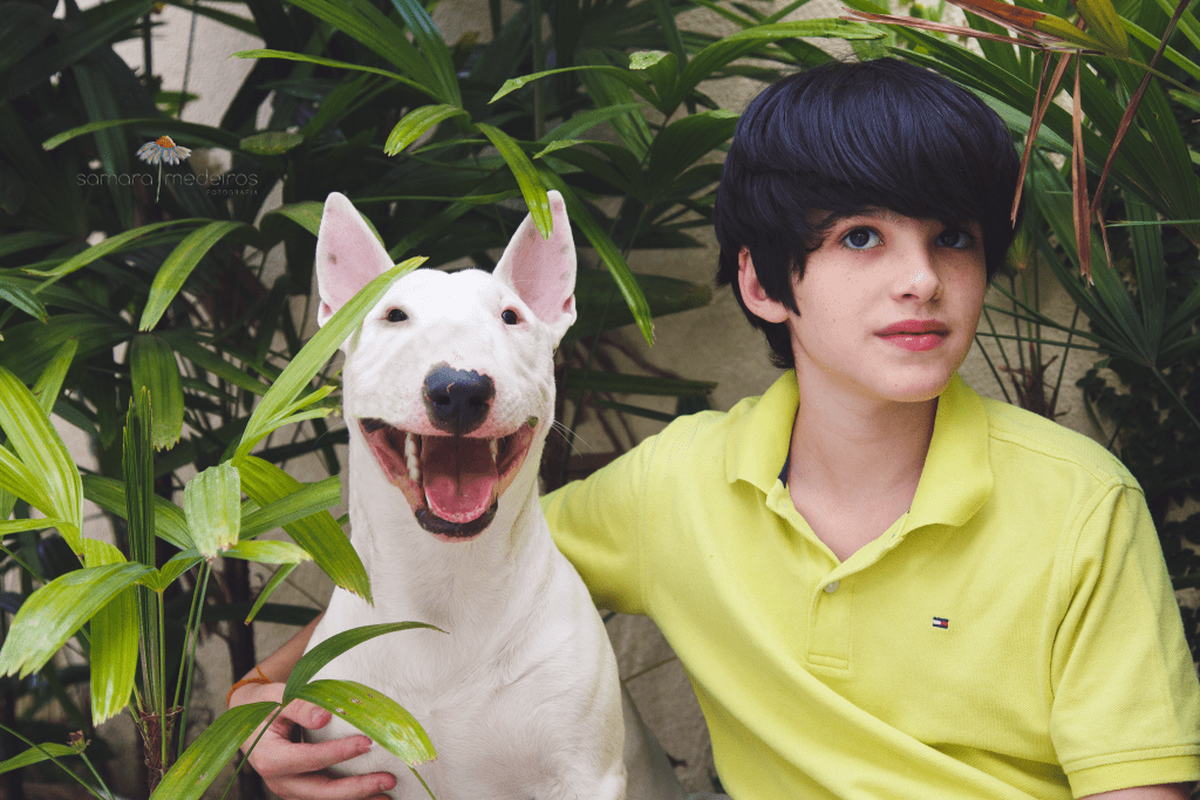 Criança e bull terrier sentado em meio às plantas do jardim, ambos olhando para a câmera, a criança séria e o bull com a boca aberta como se estivesse sorrindo.