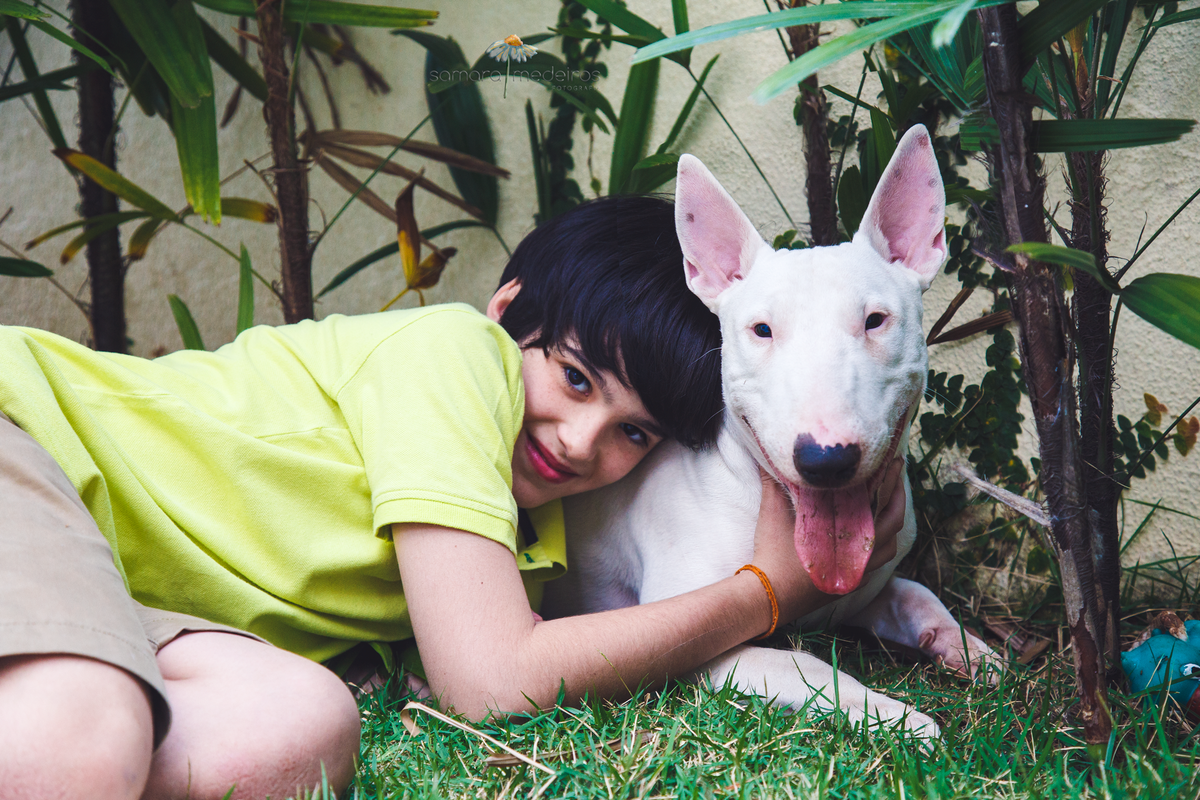 Criança e bull terrier deitados na grama do jardim abraçados, ambos olhando para a foto e sorrindo.