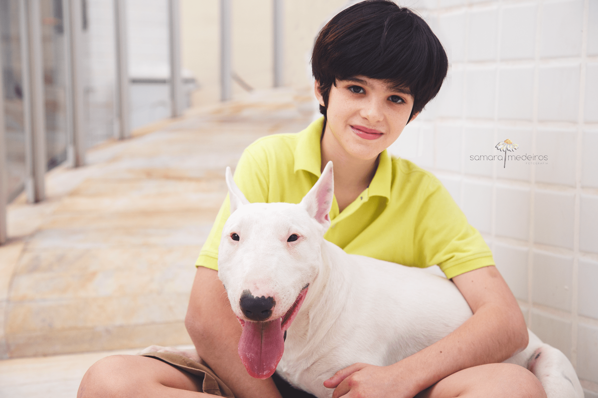 Criança com o cachorro da família, um bull terrier, no colo. Ambos olham para a câmera, a criança sorrindo e o cão com a língua para fora.