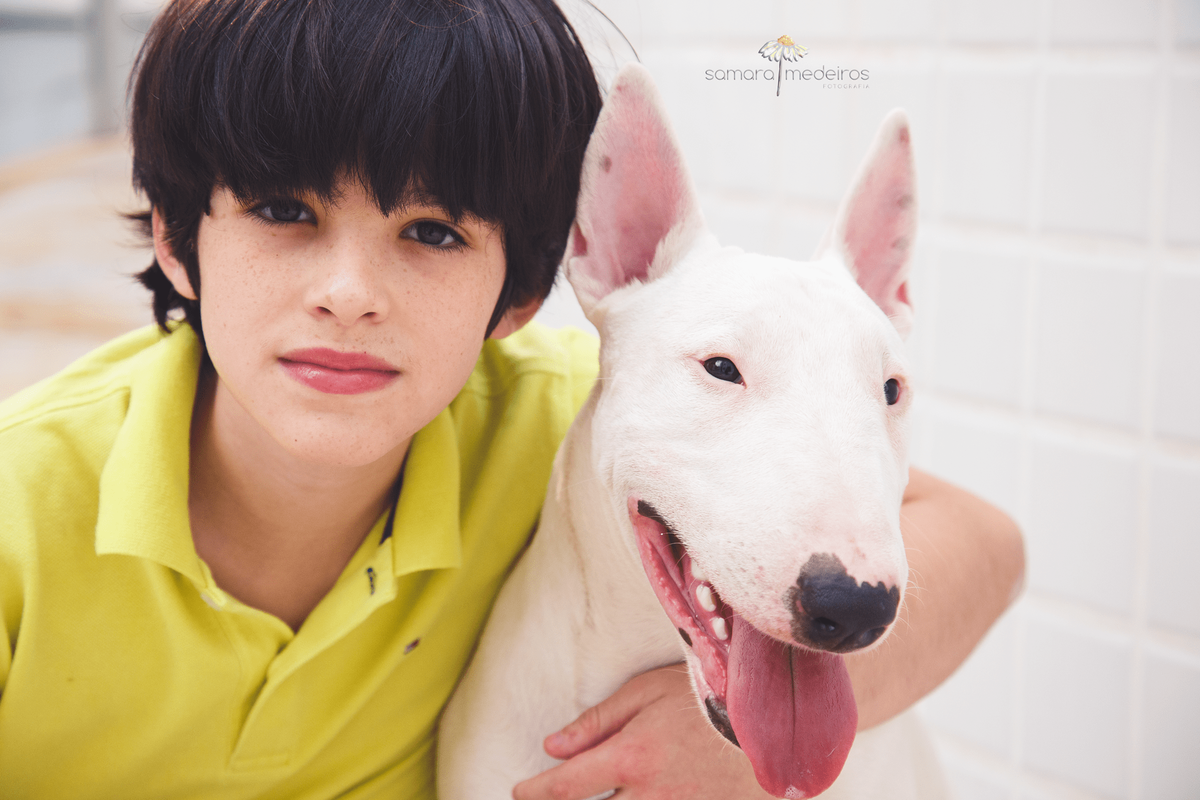 Criança abraça do pescoço do bull terrier da família e olha sério para a foto, enquanto o cachorro está com a língua para fora.