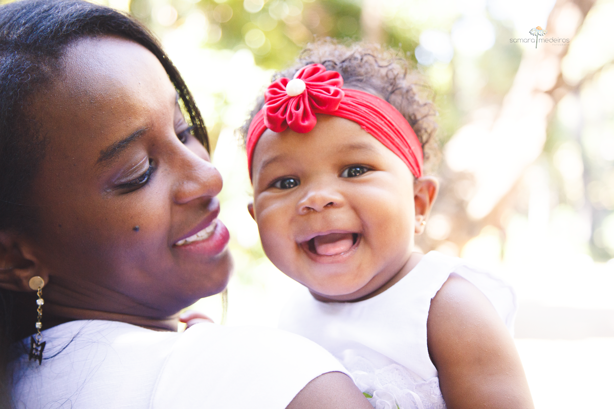 Mãe e filha em primeiro plano com árvores ao fundo. A bebê olha para a câmera e sorri e a mãe olha para a filha.