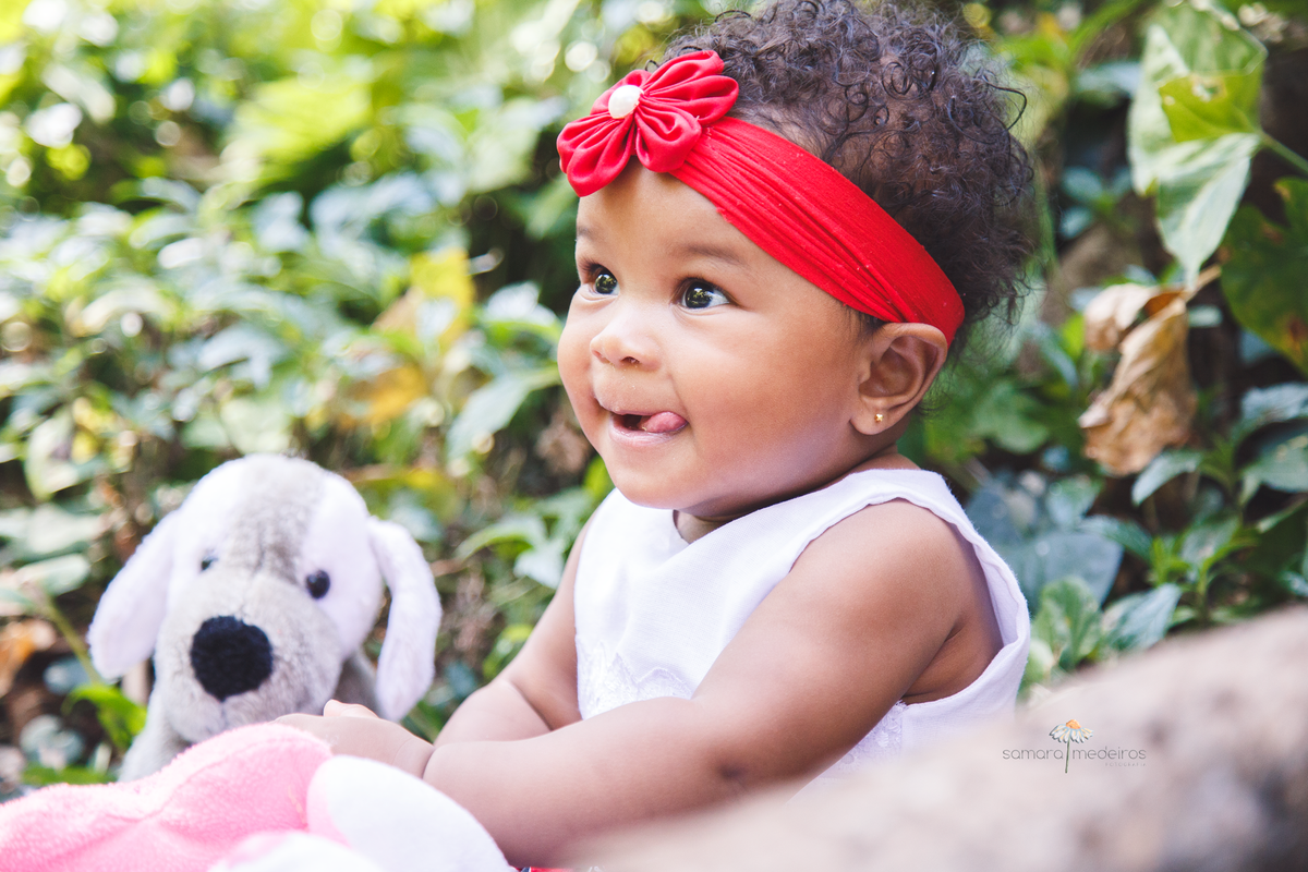 Bebê sentada na grama de um parque junto a seus ursinhos de pelúcia, ela sorri e faz uma careta.