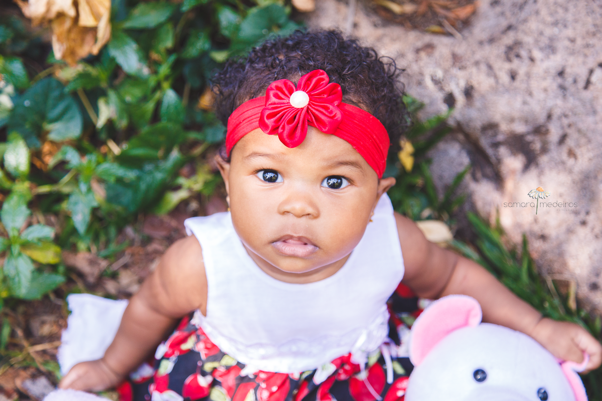 Visão de cima de uma bebê de 1 ano que está sentada na grama de um parque em Belo Horizonte. Ela olha para cima, diretamente para a câmera.