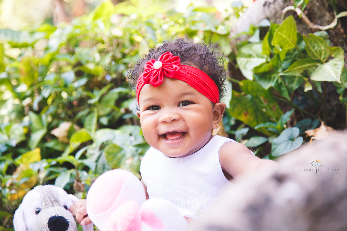 Bebê de 1 ano sentada na grama em um parque, olhando para a câmera e gargalhando.