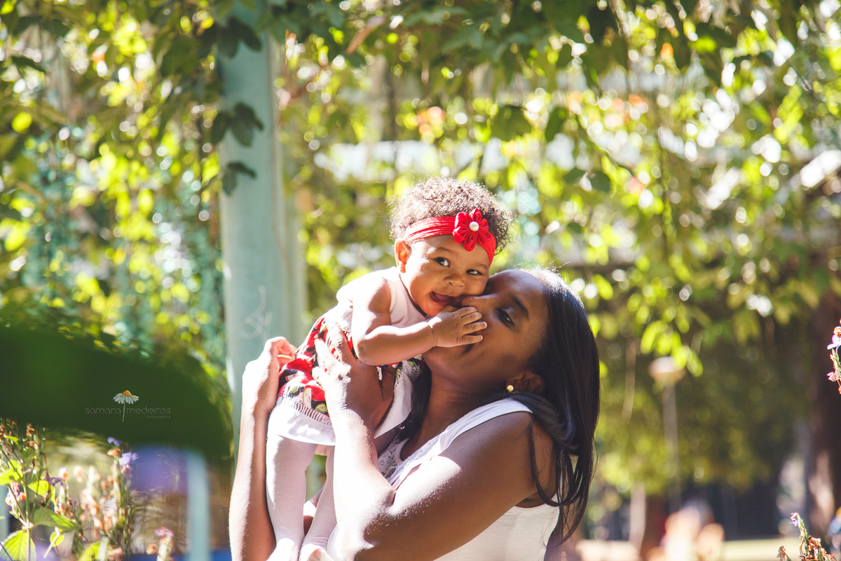 Mãe e bebê brincando em um parque de Belo Horizonte. A mãe abraça a bebê e dá um beijo, enquanto a bebê sorri.