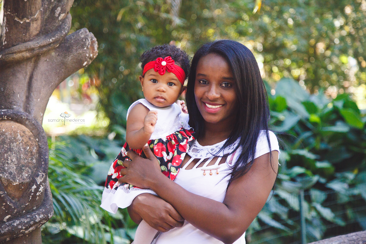 Mãe com a filha de 1 ano em um parque durante sessão de fotos de família. Ambas olham para a câmera, a bebê séria e a mãe sorrindo.