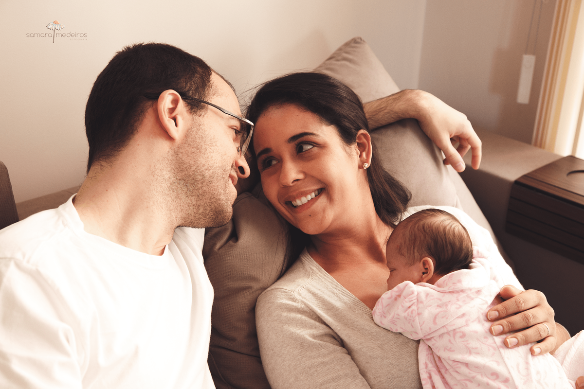 Pai e mãe sentados no sofá da casa deles se observando e sorrindo, enquanto a bebê recém nascida está deitada no colo da mãe dormindo.