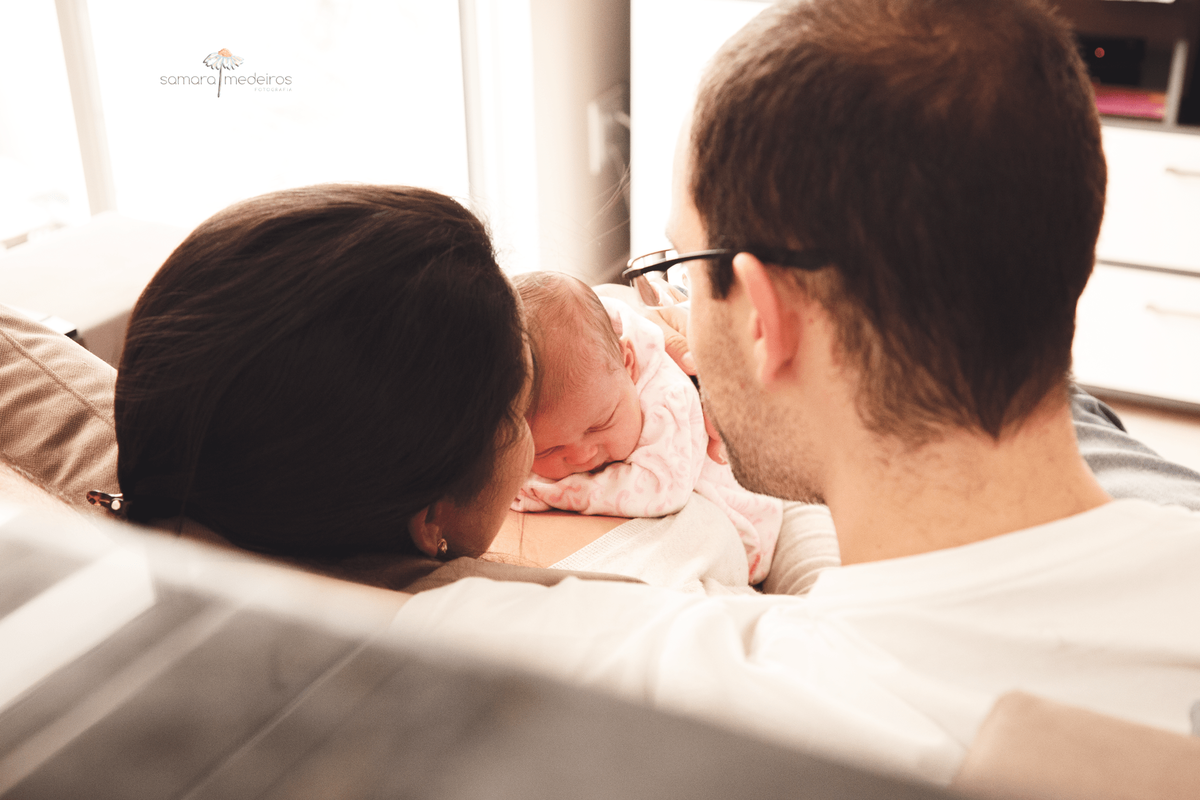 Pai, mãe e bebê recém nascido no sofá de casa, interagindo e aproveitando o momento juntos, vistos de um ângulo por trás, no qual o rosto da bebê fica em evidência e os pais estão de costas.