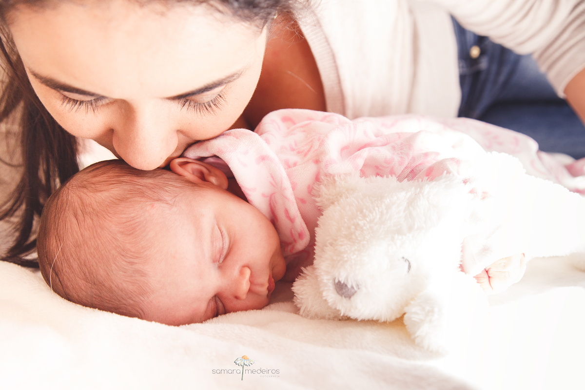 Bebê deitada na cama ao lado de um ursinho de pelúcia e a mãe cheira sua cabeça.