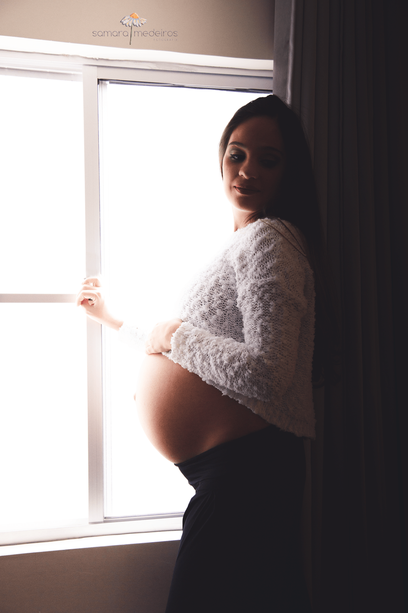 Foto com muita luz e sombras devido ao alto contraste entre a luz que entra pela janela e ilumina uma gestante, que tem a mão apoiada na barriga, e o ambiente interior escuro.