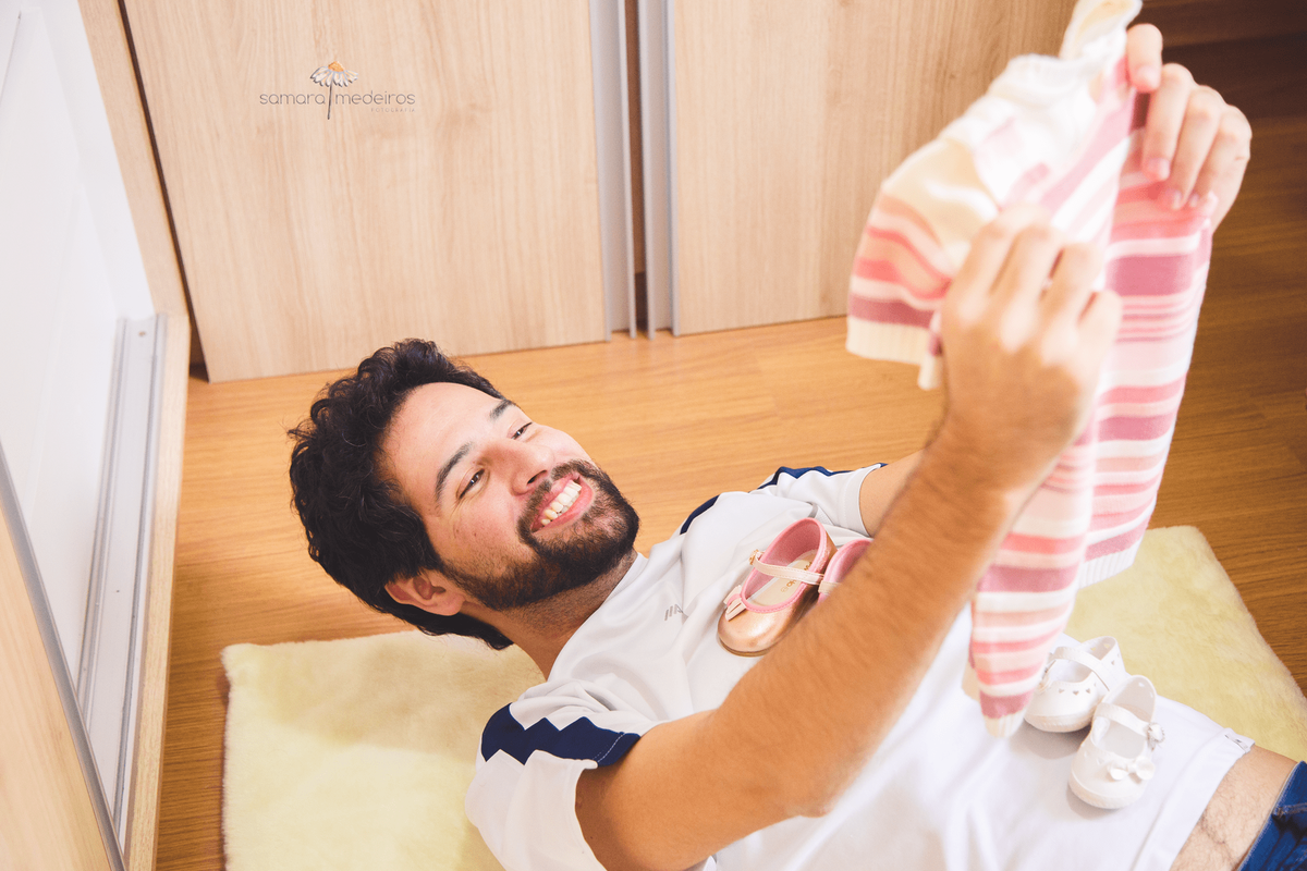 Pai deitado no chão sorrindo e segurando uma roupinha de neném sonhando com o dia que sua filha irá usar a roupinha.