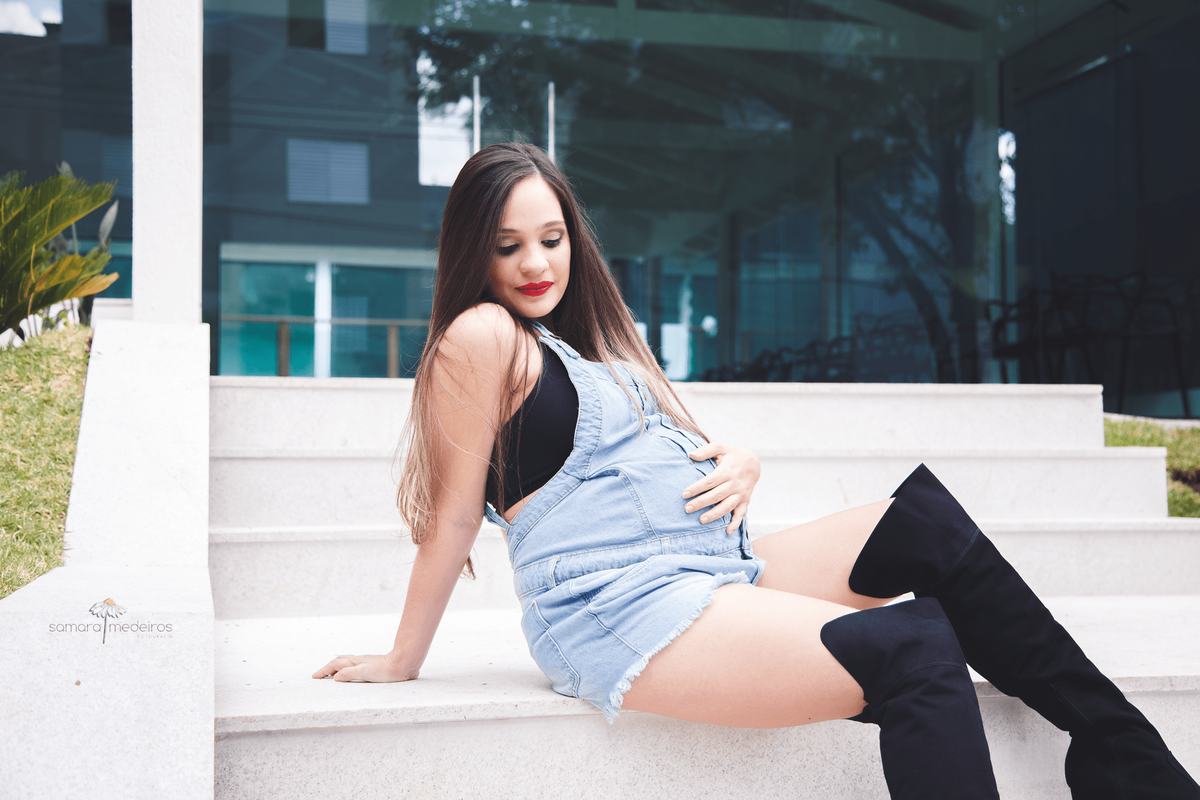 Mãe gestante sentada na escadaria do prédio, olhando para baixo e com uma mão sobre a barriga e a outra apoiando no chão.