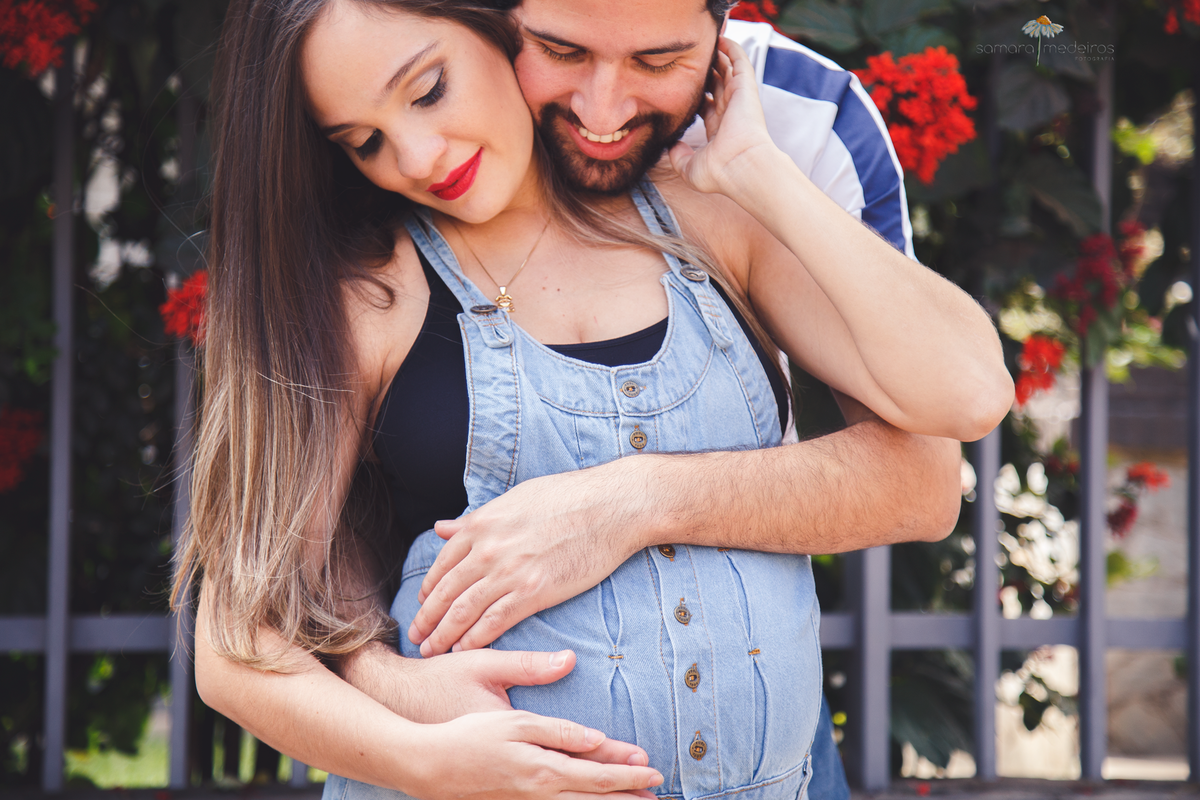 Casal abraçado, o homem atrás da mulher, ambos olhando para a barriga e fazendo carinho no bebê que está por nascer.