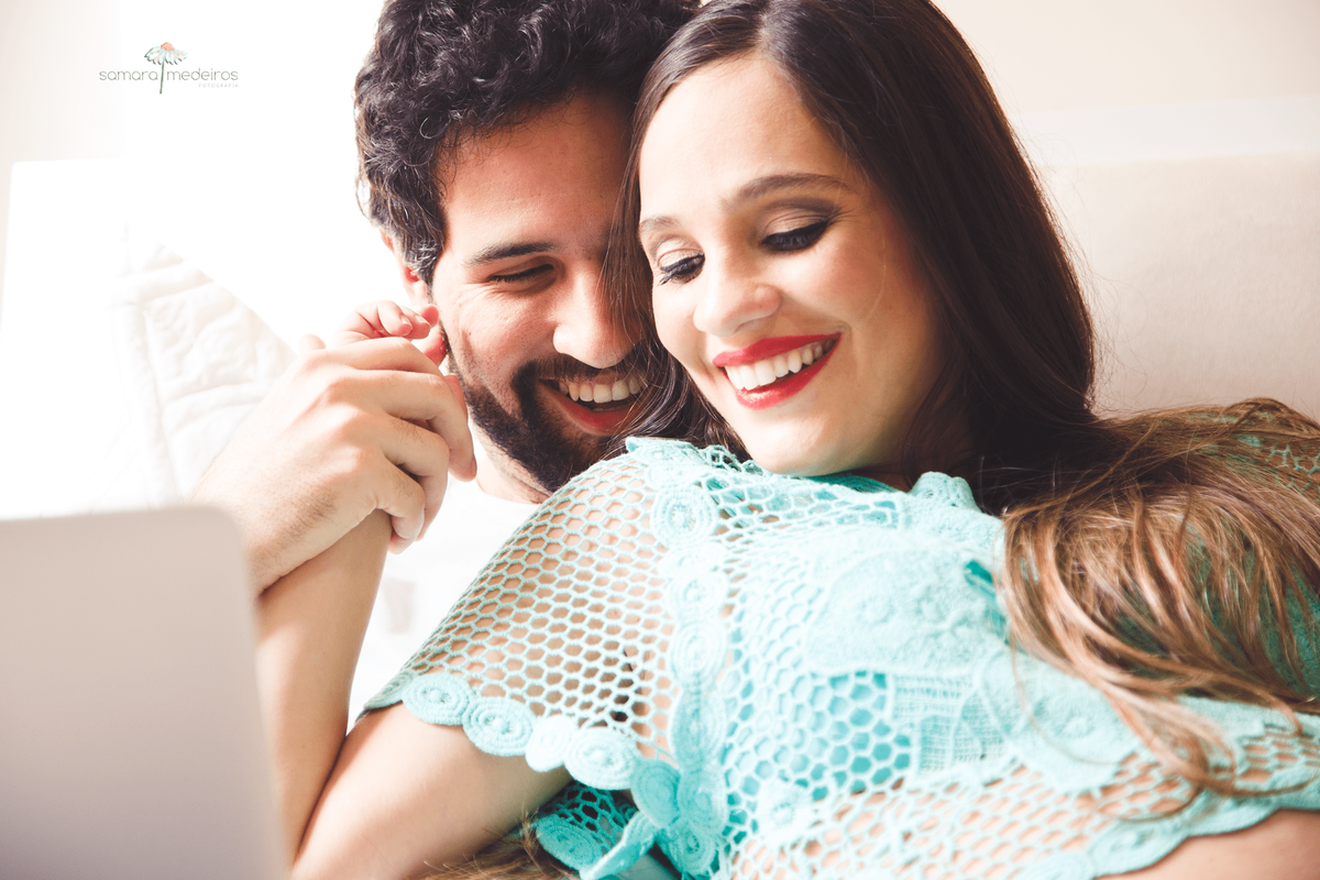 Casal sentado na cama, se abraçando e sorrindo, de mãos dadas.