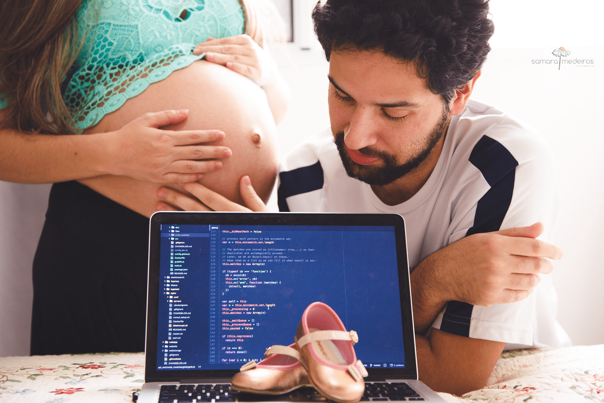 Computador no primeiro plano com um sapatinho de menina em cima do teclado e o casal ao fundo, a mãe apenas com a barriga aparecendo na foto e o pai acariciando a barriga e olhando pala a tela do computador.