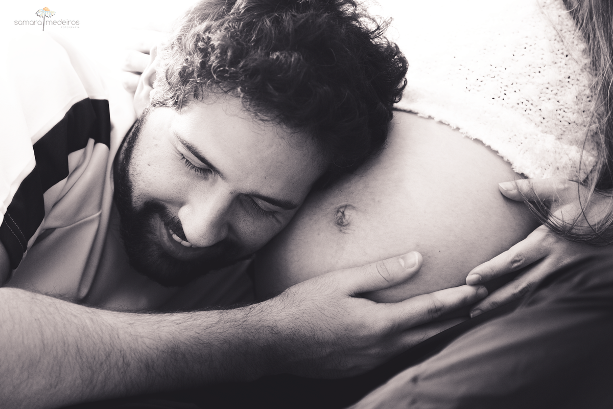 Pai sorrindo e abraçando a barriga da mãe gestante e ouvindo os sons que a bebê está fazendo lá dentro.