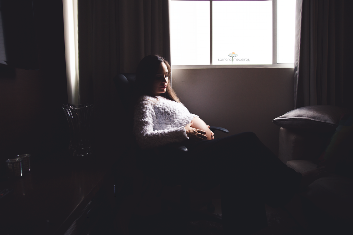 Mãe grávida sentada em uma poltrona, acariciando a barriga, lateralmente a uma janela, que deixa entrar muita luz, criando um contraste com o ambiente escuro.