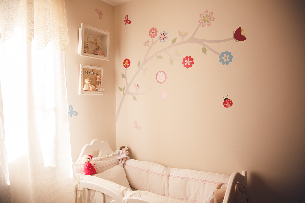 Quartinho da bebê que está para chegar, com um pedaço do berço, dois quadrinhos na parede e um papel de parede de árvore com florzinhas.