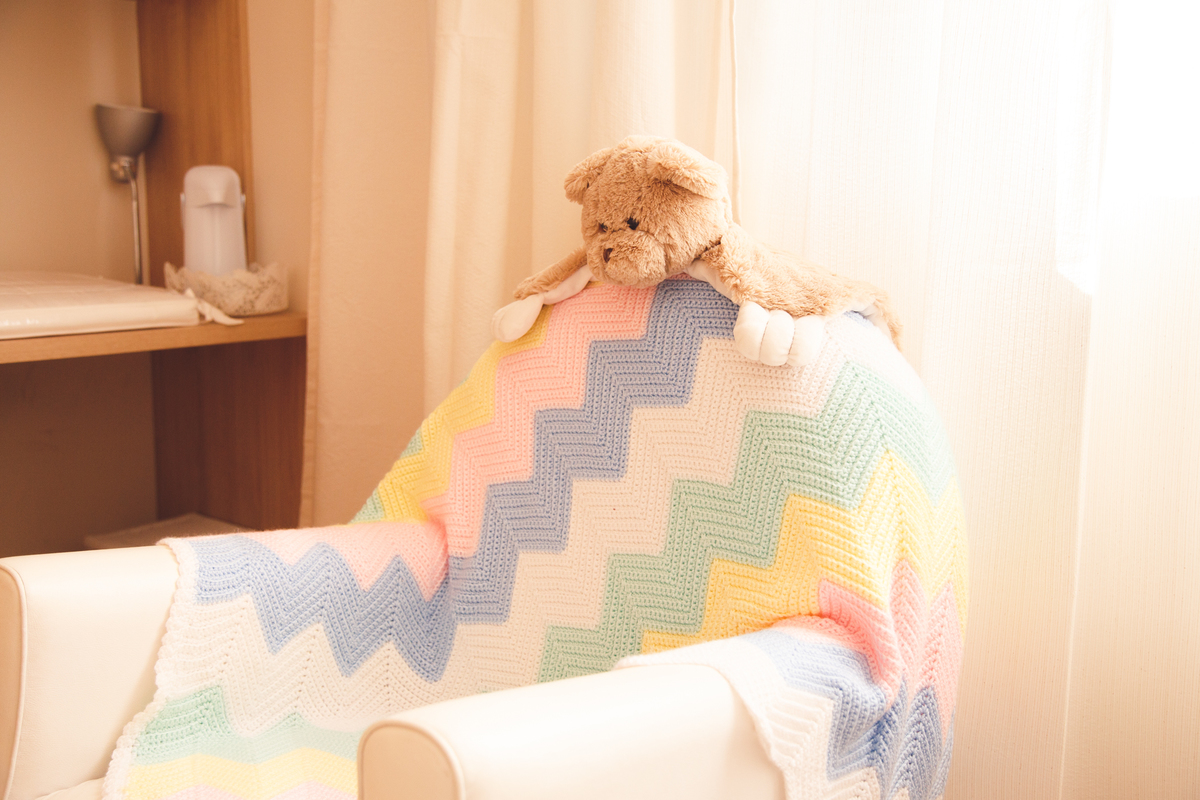 Detalhe da decoração do quartinho da bebê, com uma poltrona coberta por uma manta colorida em tons pastéis e um ursinho de pelúcia em cima do encosto.